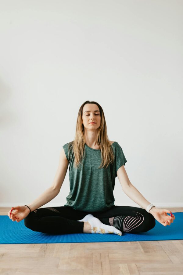 Calm slim female in sports clothes sitting with crossed legs in Padmasana pose on yoga mat while meditating with closed eyes in flat